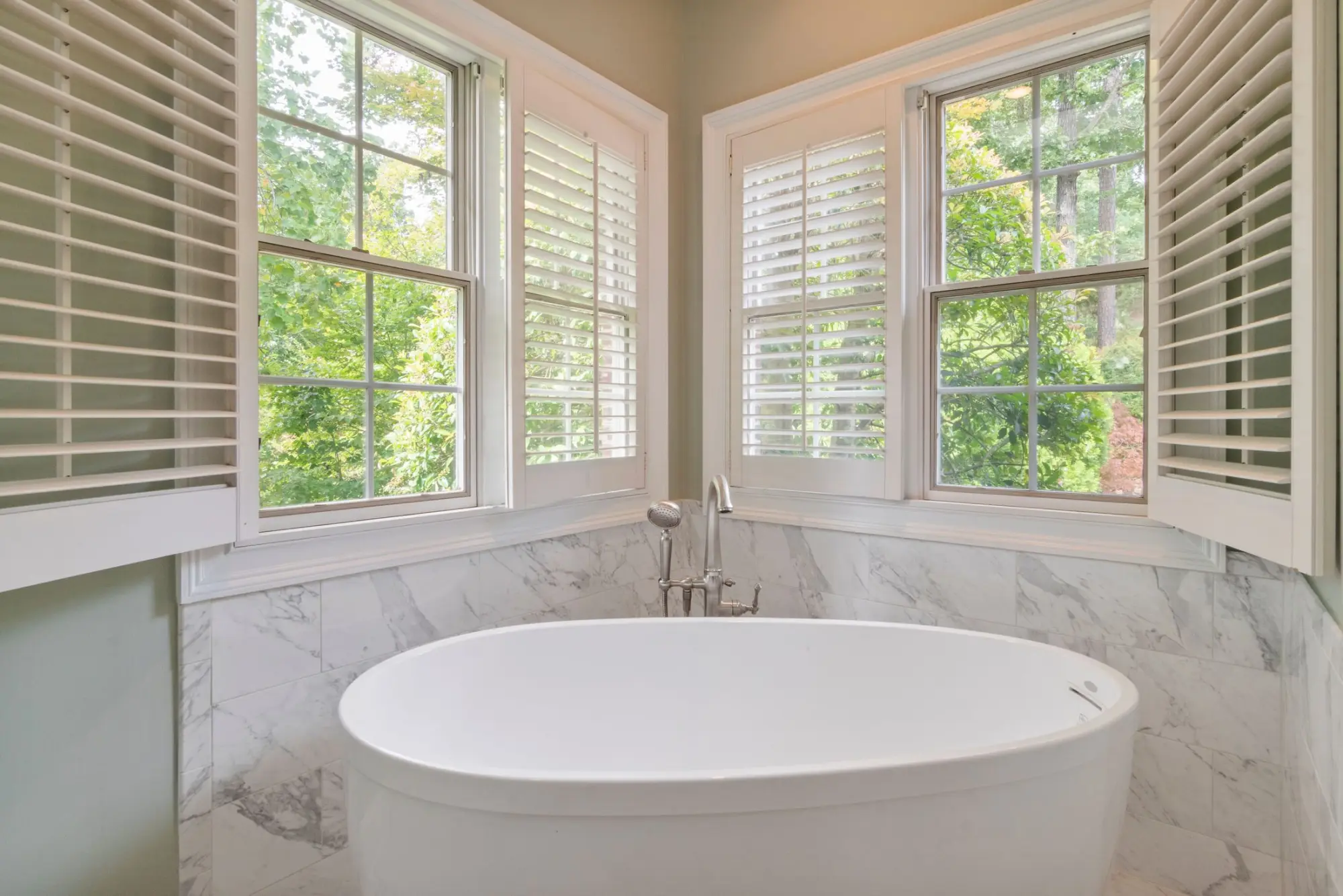 Freestanding oval tub beneath shuttered corner windows, with a marble wainscot