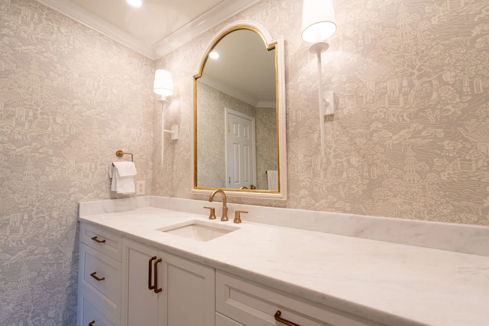 Bathroom vanity with an arched brass-framed mirror, marble top and patterned wallpaper