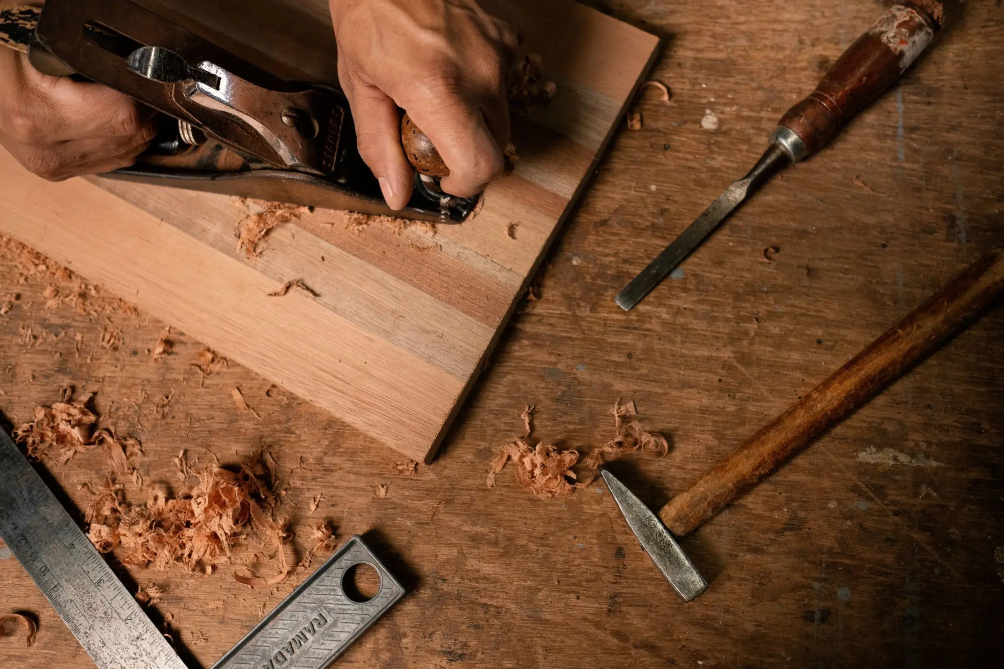 Hands guiding a hand plane across a board, with chisels and curled shavings on the bench