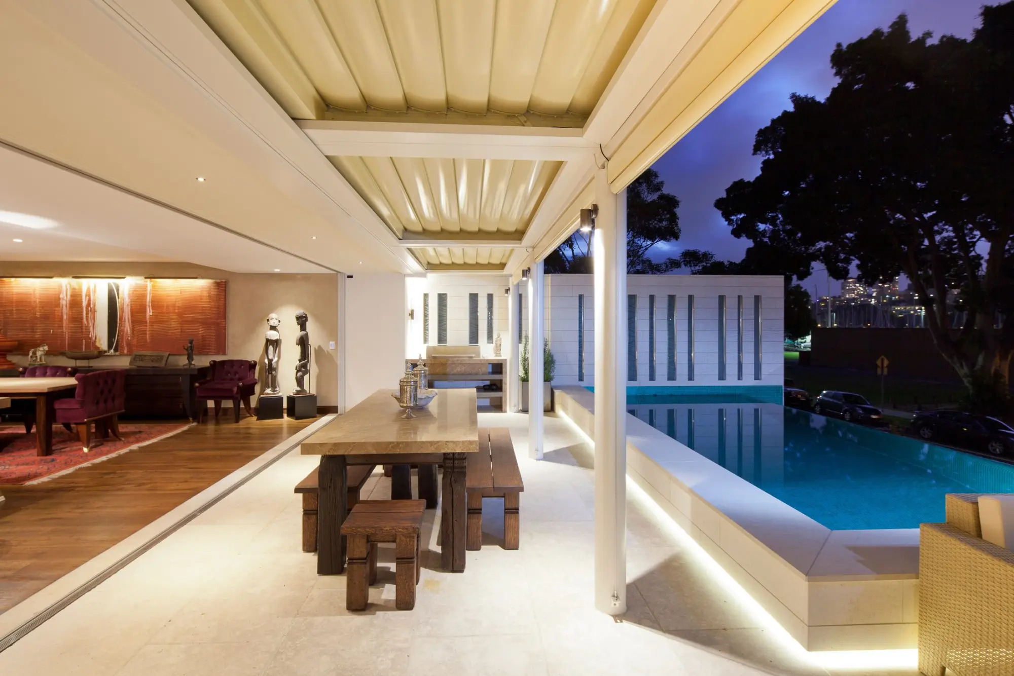 Covered patio with a timber dining table beside a lit pool at dusk