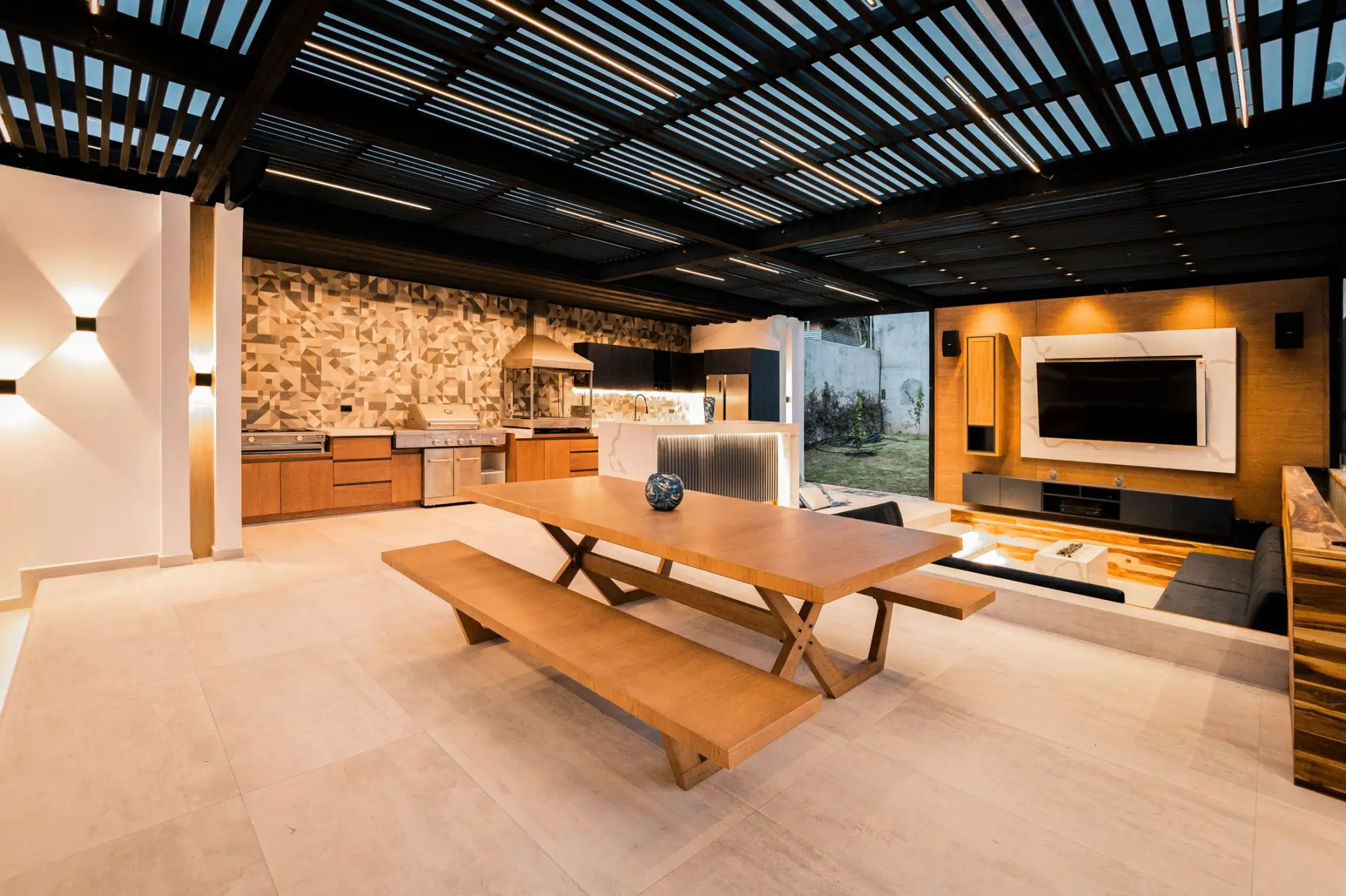 Outdoor kitchen and dining area beneath a slatted pergola, with a stone feature wall and a timber picnic table